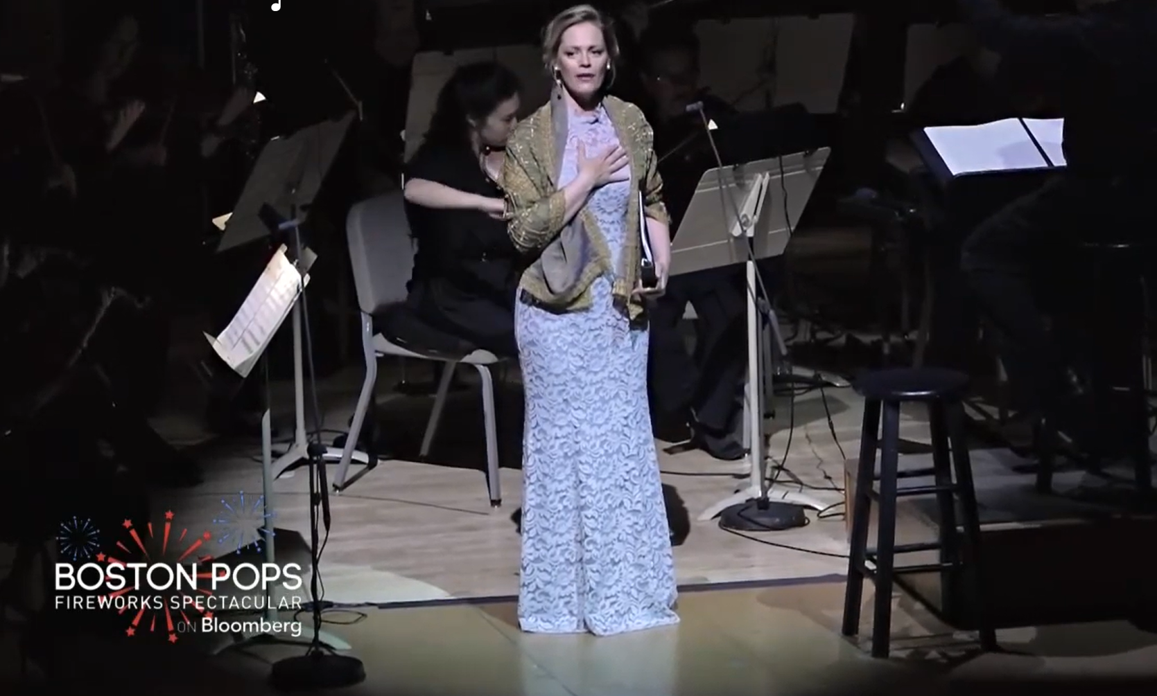A woman in a light blue dress and shawl sings on stage with musicians behind her.